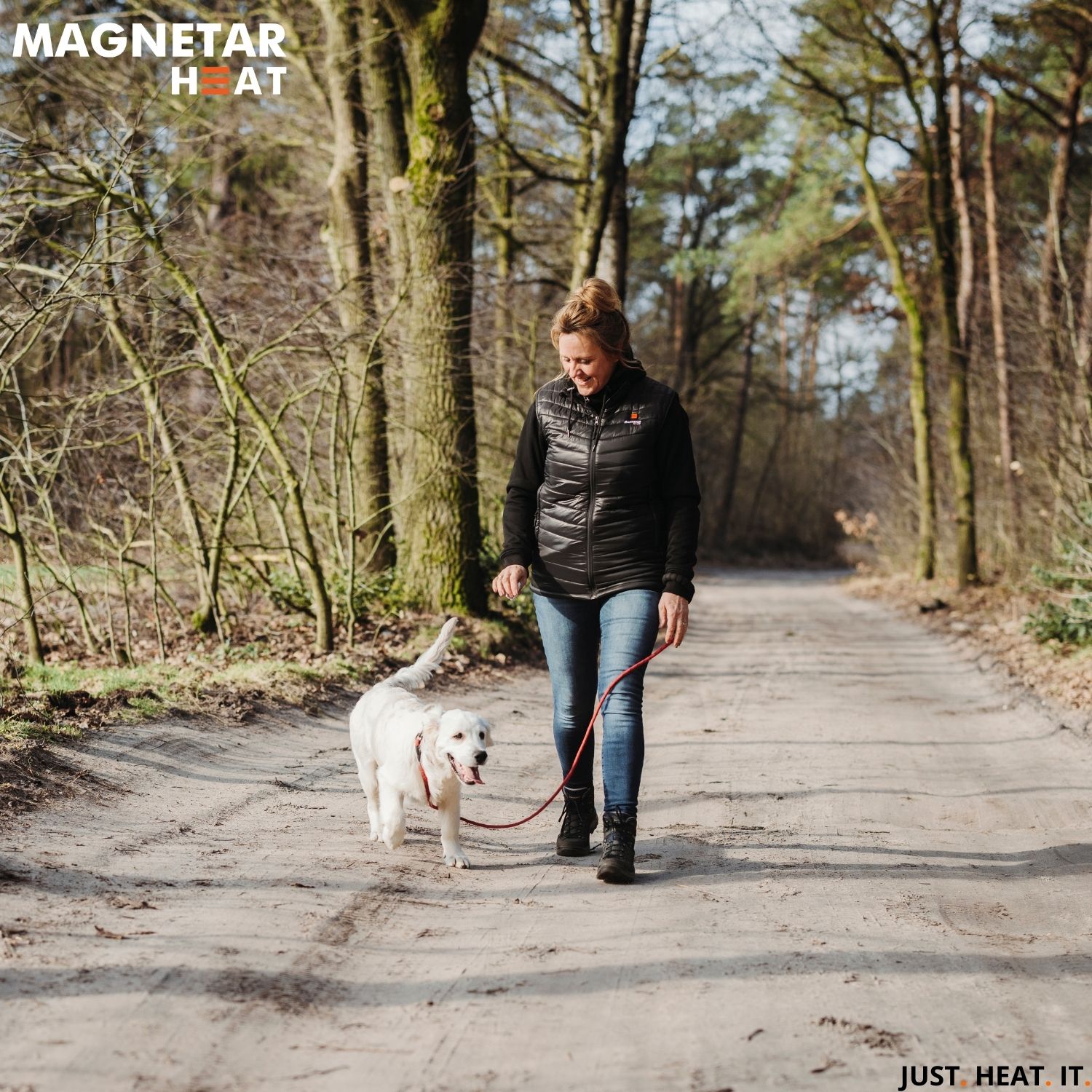 Frau trägt beheizbare Magnetar Heat Weste beim Spaziergang mit Hund im Wald, ideal für Outdoor-Aktivitäten an kühlen Tagen.